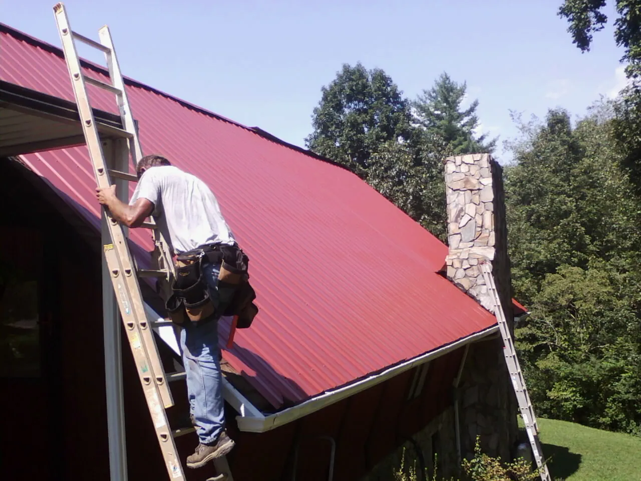 Roofer on a ladder beside a red metal roof and stone chimney — Soffit & Fascia Repair by Roofing Brothers in Drury